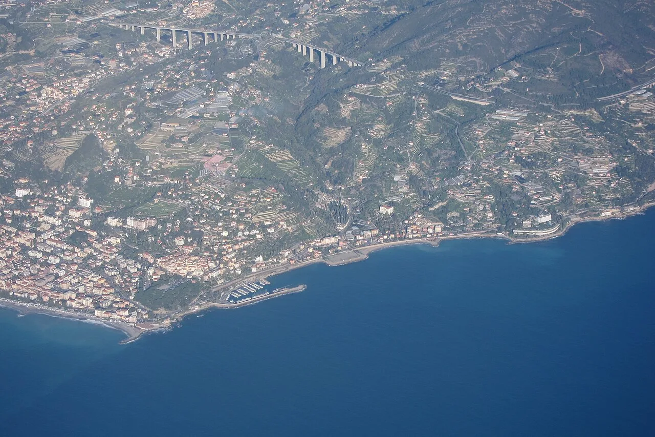 Vista aerea di Bordighera: il centro storico su promontorio, il lungomare alberato e la spiaggia sabbiosa della Riviera dei Fiori