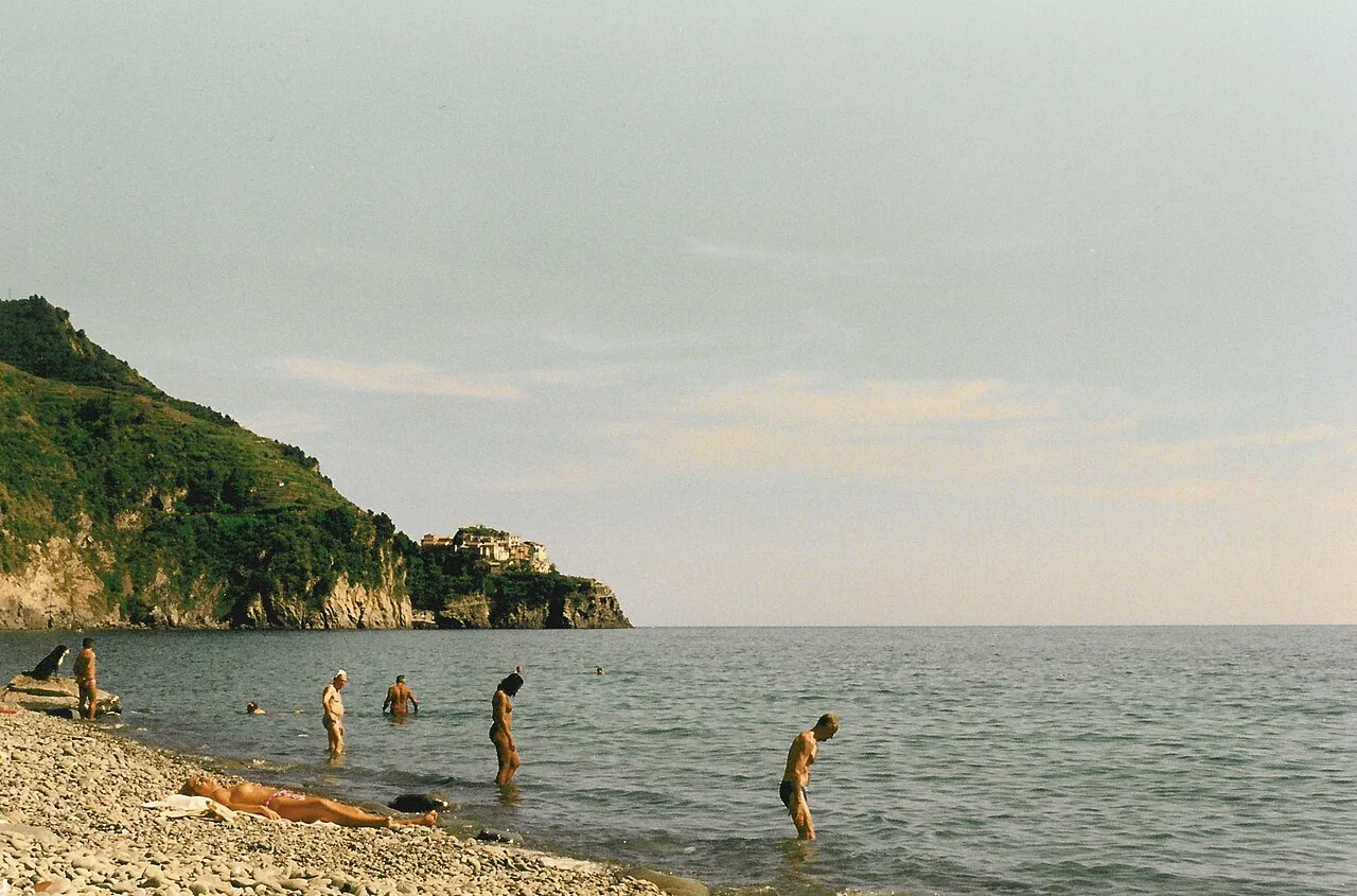 Vista dalla spiaggia di Corniglia verso Manarola: costa a picco, acqua verde e il borgo aggrappato alla roccia in lontananza