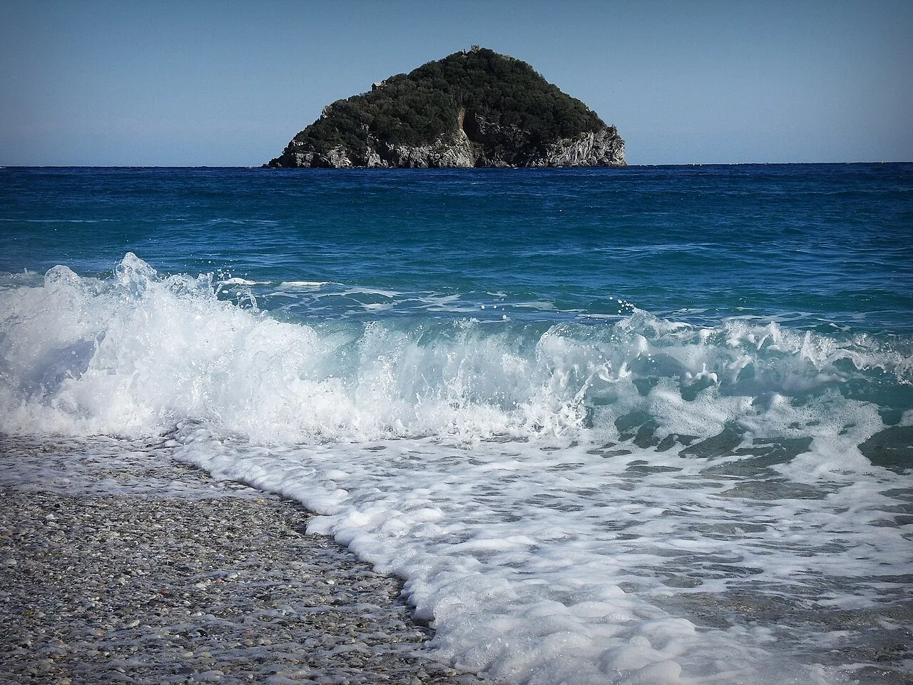 Un'onda sulla riva di ghiaia della spiaggia di Bergeggi, con l'Isola di fronte
