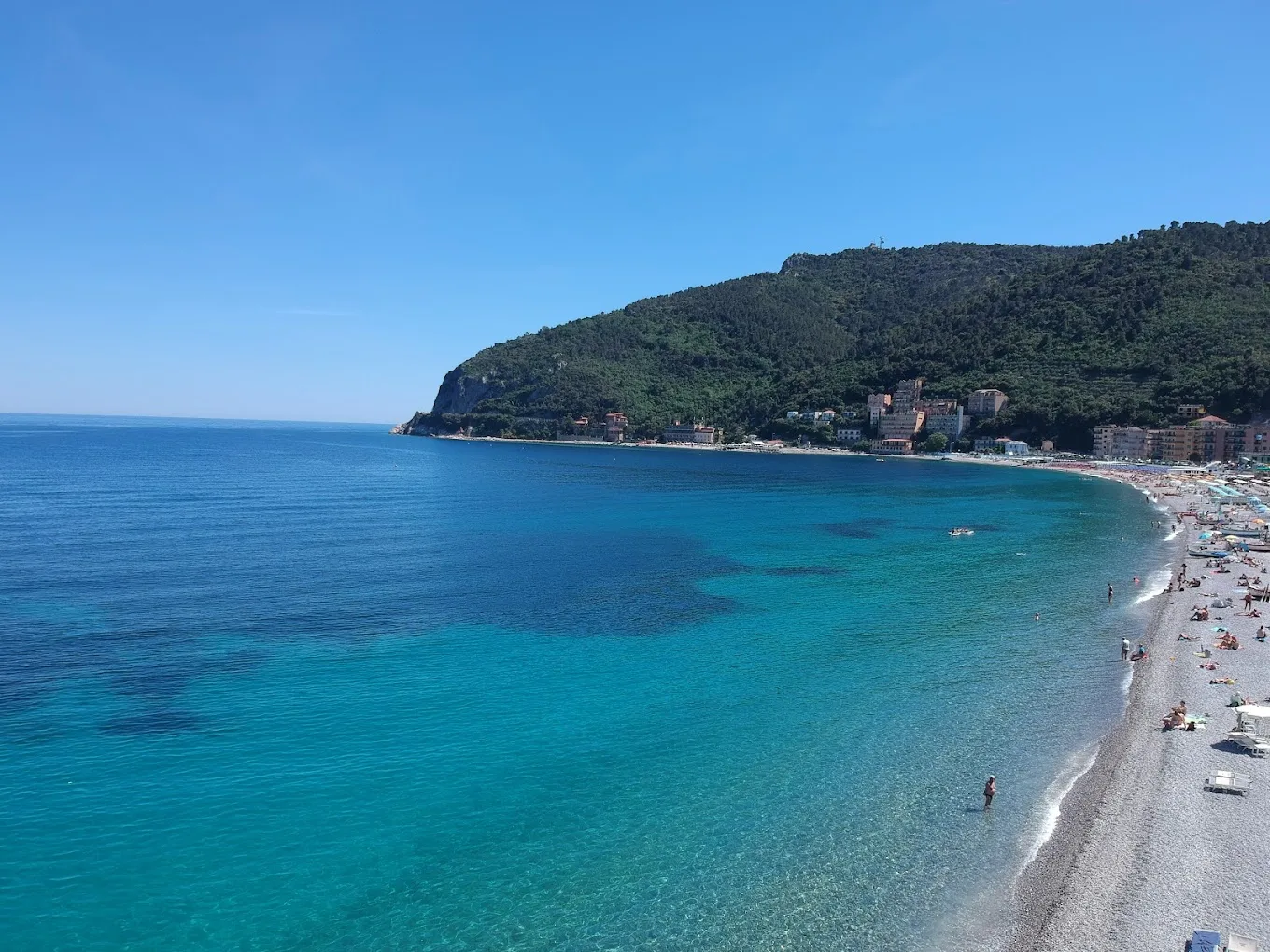 La baia di Noli con l'acqua turchese, la spiaggia di ghiaia e il promontorio di Capo Noli sullo sfondo